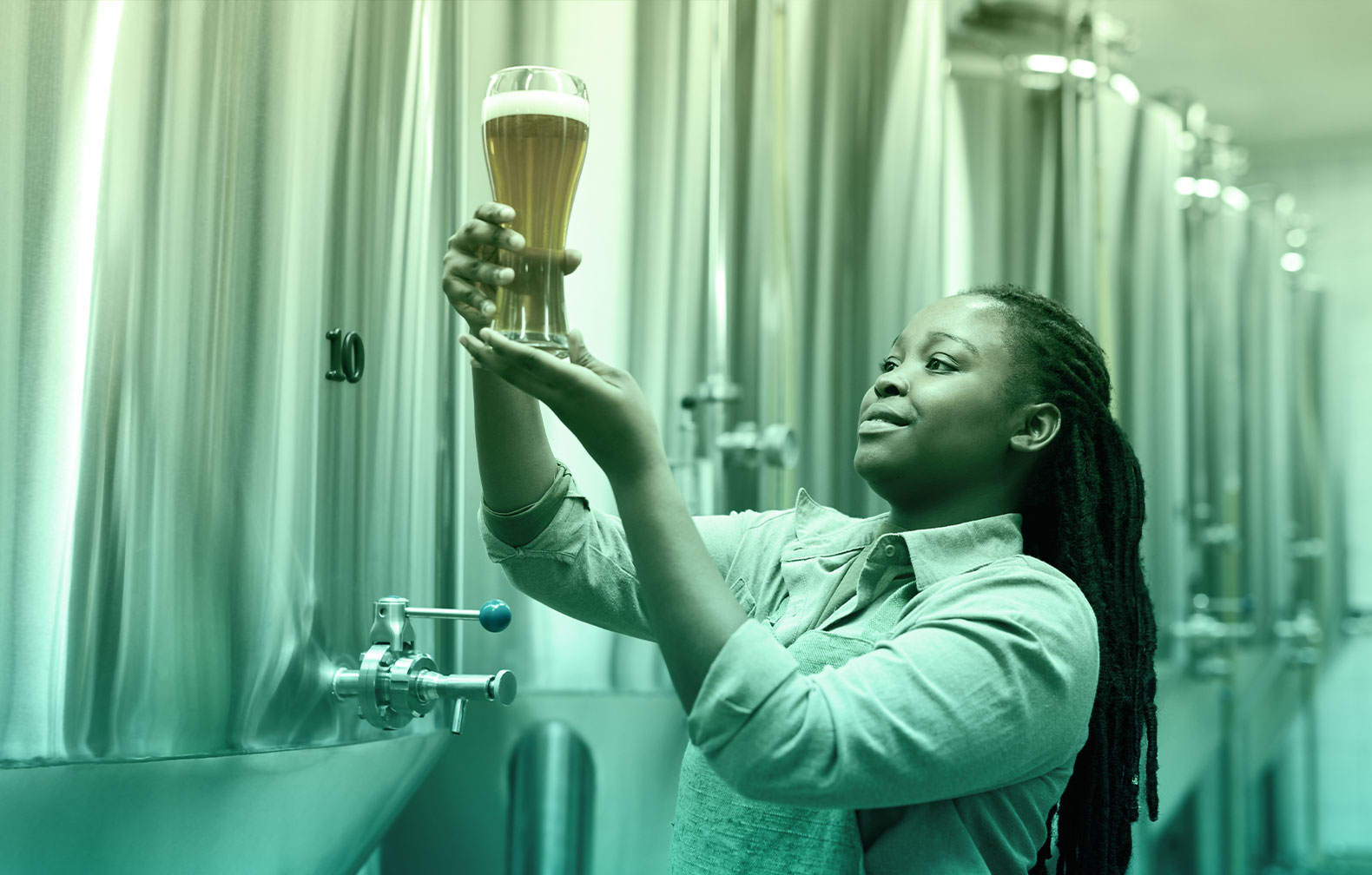 Brewey technician examining color and characteristics of recently poured beer . Fermentation vessels in background