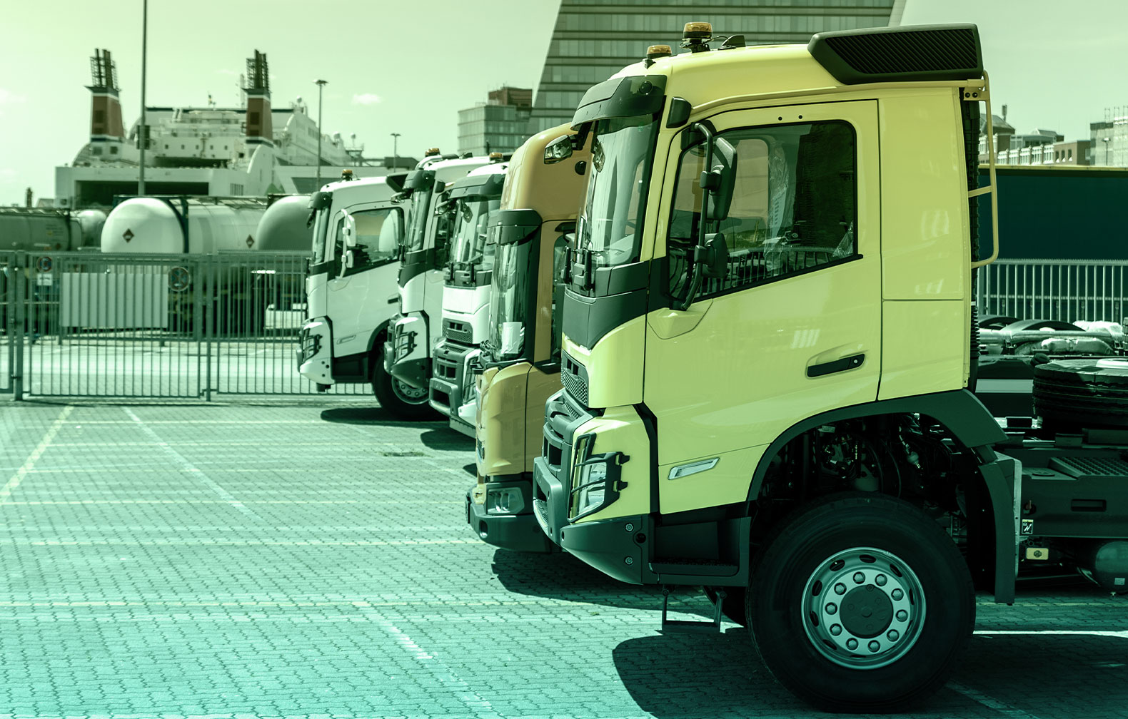 Truck fleet lined up, five visible trucks carrying no cargo, most likely in maintenance bay. In the background a large cruise-like ship can be seen