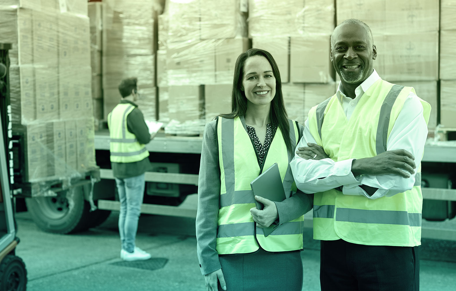In the foreground a brunette lady holding an tablet and an african american man with arms crossed. He is wearing a wedding ring. Both are wearing high visibility vests. In the background a stock auditor wearing a high visibility vest is examining cargo on a truck and annotating in a tablet. Most likely the tablet is running a CMMS app. A forklift is carrying a pallet full of boxes to load it onto the same truck.
